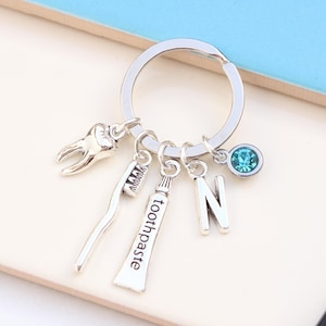 Op de afbeelding: Zilveren sleutelhanger met een tand, een tandenborstel, tandpasta en een hanger met de letter N. Een turquoise blauwe strasssteen is ook aan de sleutelhanger bevestigd.