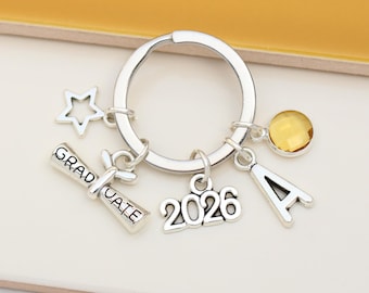 May include: A silver-toned metal keyring featuring a circular ring with several charms attached: a star outline, a diploma scroll charm engraved with the word GRADUATE, the number 2026, a letter A, and a small round yellow crystal bead.
