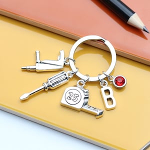 Op de afbeelding: Zilveren sleutelhanger met een rood juweel, een letter 'B', een meetlint met het nummer '25', een schroevendraaier en een boor.