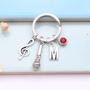 Könnte beinhalten: Silberner Schlüsselanhänger mit einem Musiknoten-Anhänger, einem Mikrofon-Anhänger, einem Buchstaben "M"-Anhänger und einem roten Strassstein-Anhänger.