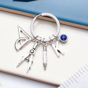 Könnte beinhalten: Silberner Schlüsselanhänger mit einem blauen Kristall-Anhänger, einem Kompass, einem Bleistift, einem Winkelmesser und einem Anhänger mit dem Buchstaben K.