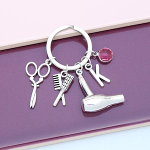May include: A silver-toned metal keychain featuring four charms: a pair of scissors, a hair comb, a hair dryer, and the letter K, accompanied by a small, round, faceted pink crystal bead attached to the circular split ring connector.