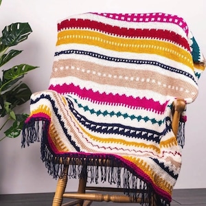 Könnte beinhalten: Eine bunte gestreifte Häkeldecke mit Fransen auf einem Holz-Schaukelstuhl. Die Decke hat verschiedene Farben, darunter Rot, Pink, Gelb, Blau und Grün.