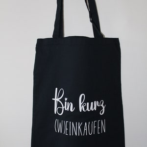 Könnte beinhalten: Schwarze Tragetasche mit weißem Text "Bin kurz (W)einkaufen". Die Tasche hat zwei schwarze Träger und hängt vor einem weißen Hintergrund. Der Text ist in einer Schreibschrift geschrieben.