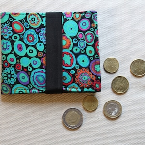 Könnte beinhalten: Eine kleine schwarze Geldbörse mit einem bunten Stoffbezug, der ein Muster aus Kreisen und Wirbeln in Blautönen, Grün, Rot und Orange zeigt. Die Geldbörse hat einen schwarzen Gummibandverschluss. Um die Geldbörse herum liegen mehrere goldene Münzen.