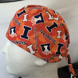 May include: An orange and navy blue patterned cap featuring the word "ILLINOIS" and the letter "I" repeated across the fabric. The cap is tied at the back and is displayed on a white mannequin head.