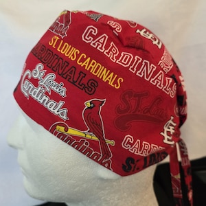 May include: Red St. Louis Cardinals scrub cap with team logos and a cardinal bird graphic. The cap features the team name in white, black, and yellow lettering. The cap is designed to be worn during medical procedures.