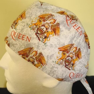 May include: A white scrub hat with a gray and white speckled pattern. The hat features a repeated pattern of the Queen band logo, which includes a lion, a phoenix, and the word "Queen" in red.