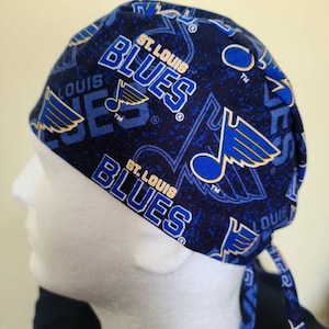 May include: A blue and gold surgical scrub hat with a repeating pattern of the St. Louis Blues logo, a musical note, and the team's name. The hat has a tie on the back.