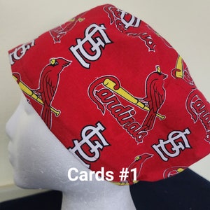 May include: Red fabric cap with a repeating pattern of the St. Louis Cardinals logo, including the team name, a cardinal bird, and a baseball bat. The text "Cards #1" is printed on the front.