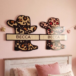 Puede incluir: Dos decoraciones de pared de madera con forma de sombreros de vaquero y botas con estampados de leopardo. Cada pieza cuenta con una placa rectangular horizontal con el nombre BECCA impreso en letras mayúsculas negras. La pieza de la izquierda tiene manchas marrones y negras, mientras que la de la derecha tiene manchas rosas y negras.