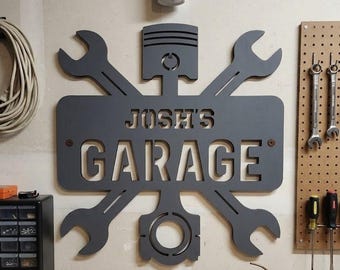 May include: A gray metal garage sign with the text "Josh's Garage" cut out. The sign features a piston, wrenches, and other mechanical elements, suitable for a garage or workshop.