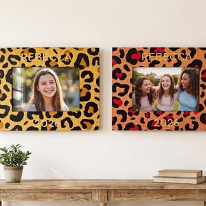 Puede incluir: Dos marcos de fotos rectangulares con estampado de leopardo. El marco izquierdo es amarillo con manchas negras, y el derecho es naranja con manchas negras y rojas. Ambos marcos muestran el nombre REBECCA y el año 2026 en letras doradas.