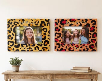 May include: Two rectangular picture frames featuring a leopard print pattern. The left frame is yellow with black spots, and the right frame is orange with black and red spots. Both frames display the name REBECCA and the year 2026 in gold lettering.