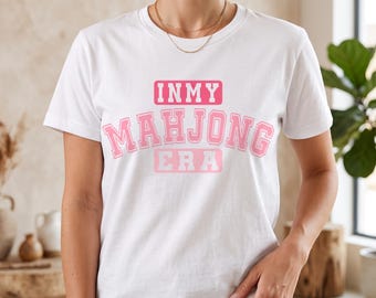 May include: A white short-sleeved t-shirt featuring the text IN MY MAHJONG ERA printed in a pink collegiate-style font across the chest. The design consists of the words IN MY in a pink rectangle, MAHJONG in large pink letters, and ERA in a pink box.