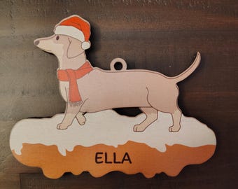 May include: A flat wooden ornament shaped like a dachshund wearing a red Santa hat and a matching red scarf. The dog stands on a white snow-covered base with the name ELLA printed in black letters. A small circular hole is at the top for hanging.