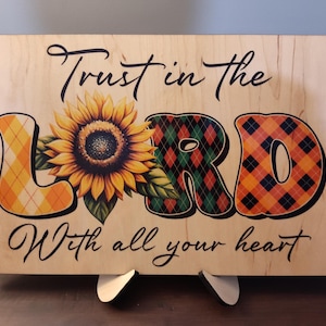 May include: Wooden sign with the text "Trust in the Lord With all your heart." The word "Lord" is decorated with a sunflower and plaid patterns in orange, green, and yellow. The sign is displayed on a wooden stand.