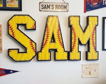 May include: Yellow wooden letters spelling "SAM" with red stitching details, resembling a baseball. The letters are mounted on a white wall, along with a sign that reads "SAM'S ROOM". Baseball-themed room decor.