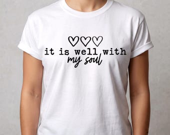 May include: A white short-sleeved t-shirt featuring three black outlined heart icons centered above the black text that reads, it is well, with my soul. The shirt has a crew neckline and a standard fit, displaying a clean and simple graphic design.