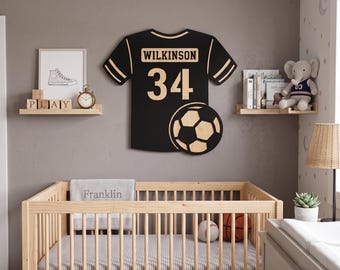 May include: A black and tan wall-mounted wooden sign shaped like a sports jersey with the name WILKINSON and the number 34 printed on it, featuring a soccer ball graphic in the lower right corner, displayed above a wooden crib in a nursery.