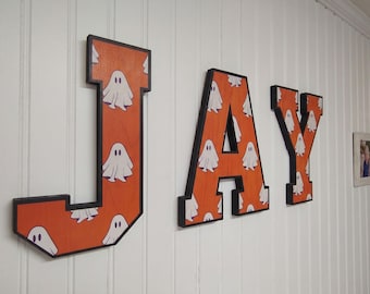 May include: Three large wooden letters spelling JAY, each featuring a vibrant orange background with a repeating pattern of white ghost illustrations. The letters have thick black edges and are mounted on a white vertical paneled wall.