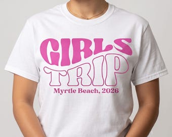 May include: A white short-sleeved t-shirt featuring the text GIRLS TRIP in large, bold, pink, wavy-style lettering across the chest. Below this, the text Myrtle Beach, 2026 is printed in a smaller, simple, dark pink serif font on the shirt.