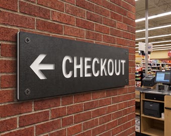 May include: A rectangular black sign with a textured surface mounted on a red brick wall. The sign features the white text "CHECKOUT" in capital letters next to a white left-pointing arrow. Four silver screws secure the sign at each corner.