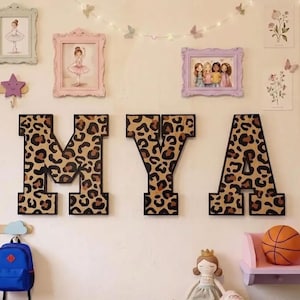 May include: Three large, block-style wall letters spelling MYA, each featuring a brown and black leopard print pattern with a solid black border. The letters are mounted on a wall alongside various framed art prints, wall hooks, and decor.