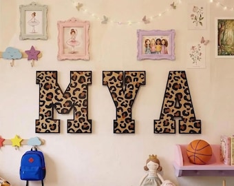 May include: Three large, block-style wall letters spelling MYA, each featuring a brown and black leopard print pattern with a solid black border. The letters are mounted on a wall alongside various framed art prints, wall hooks, and decor.