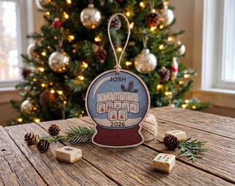 May include: A flat, snow globe-shaped wooden ornament featuring a printed illustration of mahjong tiles, the name JOSH, and the year 2026. The ornament has a light-colored wooden edge and a tan twine loop for hanging, with a dark red base design.