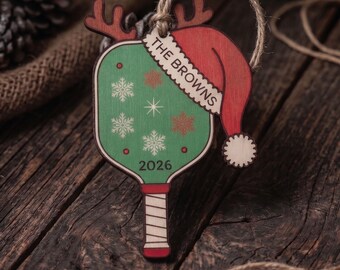 May include: A wooden pickleball paddle ornament featuring a green face with white snowflake patterns, a red Santa hat with the text THE BROWNS, brown reindeer antlers, and the year 2026 printed at the bottom, suspended by a rustic twine loop.