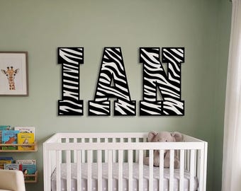 May include: Three large, block-style wall letters spelling the name IAN. Each letter features a bold, black and white zebra stripe pattern. The letters are mounted horizontally on a wall above a white crib, creating a decorative focal point.