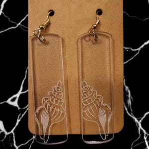May include: Clear rectangular earrings with a seashell design etched in white. The earrings have gold-colored fish hook ear wires and are displayed on a brown card against a black and white background.