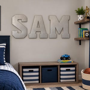 Puede incluir: Grandes letras de metal plateado que forman la palabra SAM montadas en una pared sobre una unidad de almacenamiento de madera con tres cubículos que contienen cajas de tela a rayas azules y blancas o azules sólidas.
