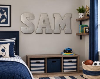 May include: Large silver-colored metal letters spelling SAM mounted on a wall above a wooden storage unit with three cubbies containing blue and white striped or solid blue fabric bins, next to a bed and corner shelves with books.