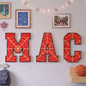 Puede incluir: Tres letras de pared grandes en estilo bloque que forman la palabra MAC, cada una con un fondo rojo vibrante con un patrón de telaraña claro y un borde negro grueso. Estas letras decorativas están montadas en la pared como adorno.