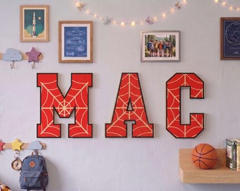 May include: Three large, block-style wall letters spelling MAC, each featuring a vibrant red background with a thin, light-colored spiderweb pattern and a bold black border. These decorative letters are mounted on a wall as a display piece.