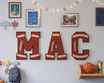 May include: Three large wooden wall letters spelling MAC, each featuring a dark red finish with tan accents and white stitching details designed to resemble the texture and appearance of an American football, mounted on a light blue wall.