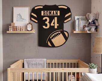 May include: A black and tan wooden wall decoration shaped like a football jersey with the name ROMAN and the number 34 printed on the front, accompanied by a graphic of a football. The piece is mounted on a wall above a wooden crib.