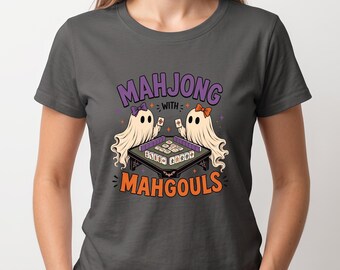 May include: A dark gray t-shirt featuring a graphic of two ghosts playing a game of mahjong. The ghosts wear small bows and are positioned around a table with tiles. The text reads "MAHJONG WITH MAHGOULS" in purple and orange lettering.
