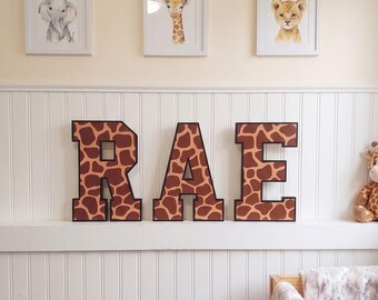 May include: Three large, block-style letters spelling RAE, each featuring a brown and tan giraffe-print pattern with black outlines. Above the letters are three white-framed prints depicting a watercolor elephant, a giraffe, and a lion cub.