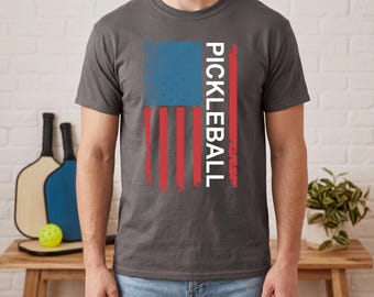 May include: A charcoal gray short-sleeve t-shirt featuring a graphic of the American flag with a distressed texture. The word PICKLEBALL is printed vertically in white capital letters over the red and white stripes of the flag design.