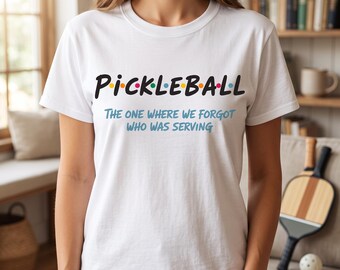 May include: A white short-sleeved t-shirt featuring the black text PICKLEBALL with colorful dots between the letters. Below this, the text reads THE ONE WHERE WE FORGOT WHO WAS SERVING in a light blue font. The shirt has a crew neckline.