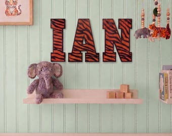 May include: Large wooden letters spelling the name IAN, each featuring a black and orange tiger stripe pattern. The letters are mounted on a light green wall above a shelf holding a gray plush elephant and three small wooden blocks.