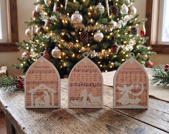 May include: Three arched wooden ornaments featuring printed sheet music and white silhouettes. The first shows a nativity scene with the text Silent Night. The second depicts shepherds and animals with Joy to the World. The third shows an angel with O Come, All Ye Faithful.