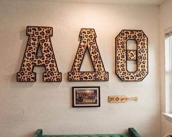 May include: Three large Greek letters, Alpha, Delta, and Theta, featuring a brown and black leopard print pattern with black borders. Below them is a framed rectangular photograph and a small wooden paddle with the letters AOK engraved on it.
