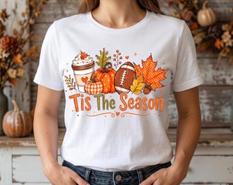 May include: A white short-sleeve t-shirt featuring a graphic print of a coffee cup, a pumpkin, a plaid pumpkin, a football, and autumn leaves. The text "Tis The Season" is printed in orange and green letters below the seasonal illustrations.