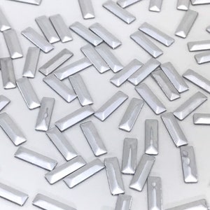 Puede incluir: Una gran cantidad de adornos rectangulares para uñas de color plateado. Los adornos están esparcidos sobre una superficie blanca.