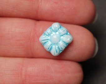 Puede incluir: Una pequeña cuenta de forma cuadrada con un patrón de relieve redondeado similar a una flor. La cuenta presenta una base de color blanco brillante con acentos azul claro vibrantes que resaltan los pétalos florales y el punto central.