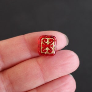 Pode incluir: Uma pequena conta de vidro vermelha de formato retangular com um motivo decorativo dourado centralizado. A conta tem um acabamento liso e brilhante com cantos arredondados, exibindo uma base vermelha translúcida vibrante com um padrão dourado simétrico.